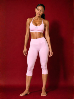 A woman modeling a light pink activewear set consisting of a sports bra and matching high-waisted capri leggings, standing against a solid dark red studio backdrop.