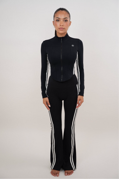 A model stands against a light gray studio background wearing a matching black athletic set with white side stripes, consisting of a long-sleeve zip-front top and high-waisted flared pants.